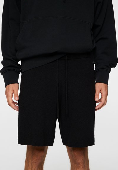 Svart hettegenser med frontlomme og snøring. Kombinert med svarte shorts, elastisk linning og strukturert stoff, som fremhever en uformell stil.