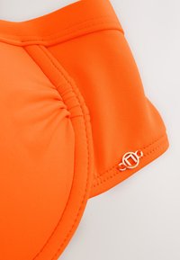 Haut de bikini rembourré orange vif avec détail froncé et petit emblème rond en argent près de l'ourlet sur fond blanc.