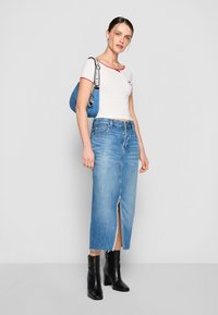 Hvit croppet topp med rød kant, blå denim maxiskjørt med frontslitt, svarte ankelstøvler, og en blå håndveske med stroppe detaljer.