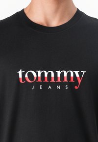 Black cotton t-shirt featuring a bold white and red "tommy" logo with "JEANS" below, classic crew neck design, smooth texture.