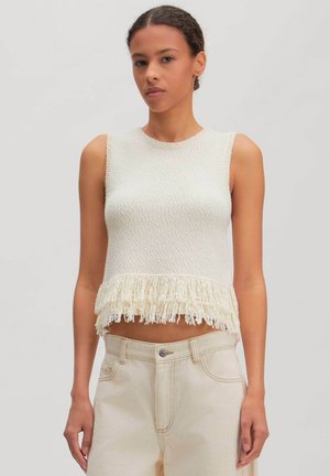 Vrouw draagt een mouwloze witte, geribbelde cropped top met franjes aan de zoom, gecombineerd met een hooggetailleerde lichtbeige broek tegen een effen achtergrond.
