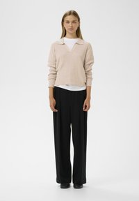 Pulover ribbed de culoare roz deschis, cu decolteu în V și guler, asortat cu pantaloni cu picioare largi, negri. Modelul stă pe un fundal simplu.