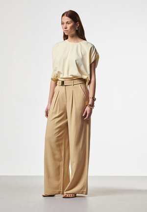 Vrouw draagt beige broek met wijde pijpen en riem, los crèmekleurig kort mouwshirt, sandalen en gouden sieraden, staand tegen een effen achtergrond.