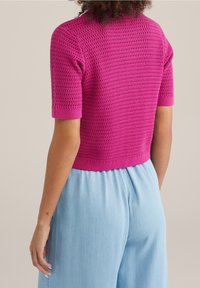 Stickad kortärmad topp i klar rosa med ett texturerat mönster och ribbad nederkant, kombinerad med ljusblå vida byxor.