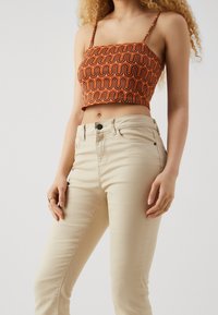 Mujer vestida con jeans de tiro alto beige y un top corto naranja con un patrón ondulado, de pie frente a un fondo claro y liso.