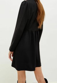 Frau mit langen kastanienbraunen Haaren, die ein schwarzes langärmliges Kleid mit gerafftem Bund trägt, steht vor einem schlichten, hellen Hintergrund.