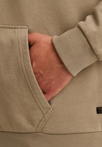 Beige sweatshirt met ribbelmanchetten en een voorkantzak; toont een hand die in de zak rust, waarbij de textuur van de stof en de stiksels worden benadrukt.