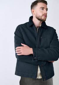 Chaqueta azul oscuro con cierre de cremallera, que presenta un cuello puntiagudo, bolsillo frontal con cremallera y detalles de costura visibles. Textura suave, diseño casual.