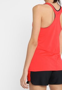 Czerwy top sportowy z designem na plecach, wykonany z gładkiego materiału, w połączeniu z czarnymi szortami z czerwonymi akcentami po bokach.