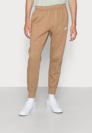 Mann trägt beige Nike Sweathose und weiße Sneaker, steht vor einem einfarbigen hellgrauen Hintergrund.