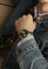 Orologio in acciaio inossidabile argento e oro con quadrante nero, funzioni cronografiche e bracciale metallico. Indossato su una mano visibile in una giacca di denim.