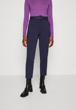 Pantaloni blu navy con vestibilità su misura, dotati di una cintura larga con fibbia rettangolare. Abbinati a un maglione aderente viola e stivaletti neri alla caviglia.