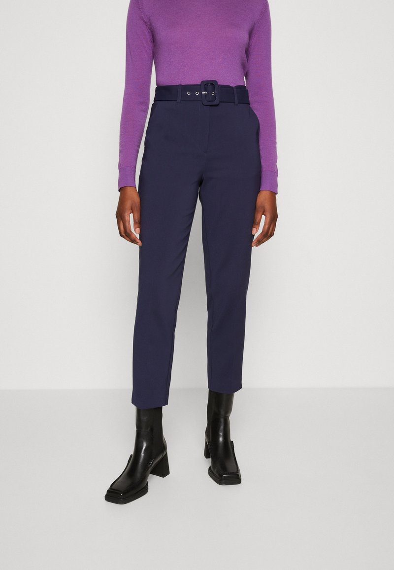 Marinblå byxor med skräddarsydd passform, utrustade med ett brett bälte med en rektangulär spänne. Tillsammans med en lila åtsittande tröja och svarta ankelskor.