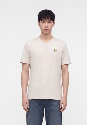 Jeune homme portant un t-shirt beige clair à col en V avec un petit logo jaune d’oiseau sur la poitrine gauche, associé à un jean bleu.