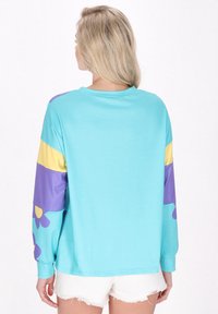 Camisa de manga larga en azul claro, con franjas moradas y amarillas y patrones florales en las mangas. Material suave, corte relajado.