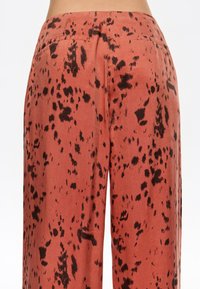 Pantalones de pierna ancha de color óxido en tejido ligero, con un patrón abstracto negro, textura suave y un detalle de bolsillo trasero.