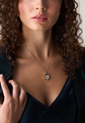 Donna con i capelli ricci che indossa un vestito scuro con scollatura a V e una collana d'oro con un pendente caratterizzato da pietre decorative trasparenti.