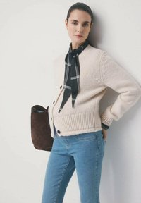 Kobieta w kremowym dzianinowym kardiganie, niebieskich dżinsach i czarnym, wzorzystym szaliku, trzymająca brązową torbę z zamszu, stojąca na tle białej ściany.