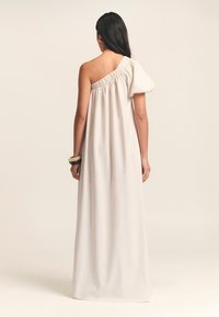 Abito maxi asimmetrico in tessuto beige chiaro, con una manica a sbuffo, corpetto elasticizzato e una silhouette fluida. Texture liscia.
