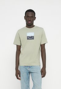 Ανοιχτοπράσινο βαμβακερό t-shirt με στρογγυλή λαιμόκοψη, με τετράγωνο γραφικό σε μπλε και ναυτικό μπλε με κείμενο, συνδυασμένο με ανοιχτό μπλε τζιν.