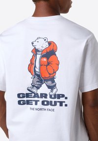 Bjørn iført en orange dunjakke, bukser og sneakers med teksten "GØR DIG KLAR. KOM UD. THE NORTH FACE" på bagsiden af en hvid t-shirt.
