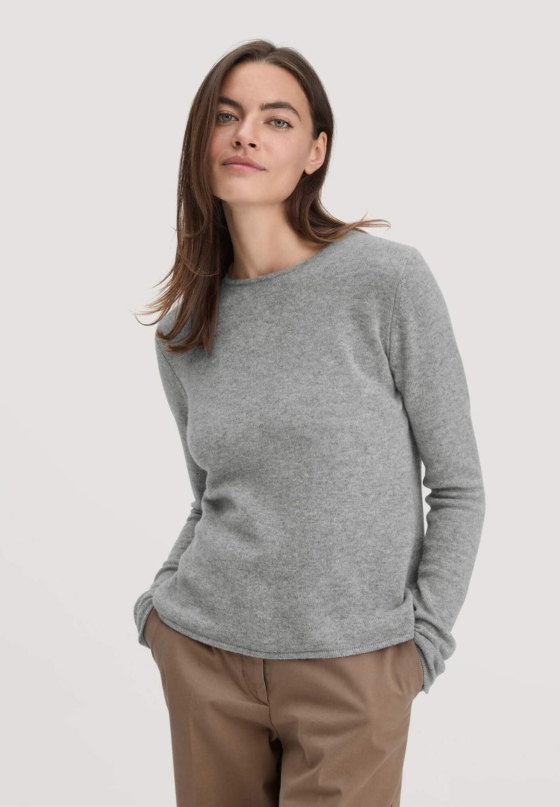 Grauer, langärmeliger Pullover mit Rundhalsausschnitt, der eine weiche Textur und gerippte Ärmelbündchen aufweist, kombiniert mit hellbraunen Hosen.