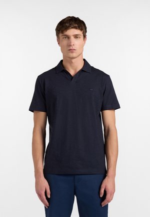 Mladý muž stojaci, oblečený v tmavomodrej polokošeli s krátkym rukávom a zladených tmavomodrých nohaviciach, smerujúci dopredu na jednoliatom bielom pozadí.