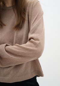 Femme aux longs cheveux bruns portant un pull beige ample par-dessus un pantalon noir, debout devant un fond clair uni, les bras croisés.