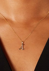 Gouden halsketting met een cursieve letter "A" hanger versierd met kleine diamanten, geplaatst tegen een getextureerde huidachtergrond.