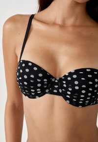 Haut de bikini noir à pois avec bonnets moulés, soutien-gorge avec armatures et bretelles ajustables ; détail de tissu froncé au niveau de la poitrine.