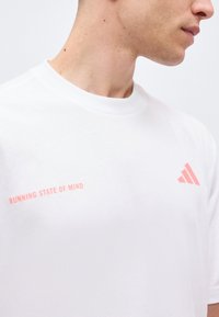 Άνδρας που φοράει λευκό t-shirt με κόκκινο κείμενο "RUNNING STATE OF MIND" και ένα κόκκινο τριγωνικό λογότυπο στο στήθος, κοιτάζοντας προς τα δεξιά.