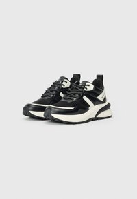 Par črno-bele športne superge z debelimi belimi podplati, črnimi vezalkami in potegovalno zanko na peti na enobarvni podlagi.