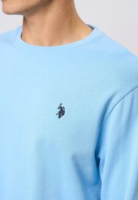 Svetlo modra bombažna majica s kapuco, ki ima krožni izrez in črn vezen logotip na prsih. Gladka tekstura in sproščen kroj.