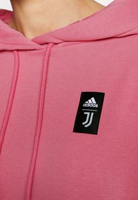 Hoodie rosa realizzato in tessuto morbido, con patch del logo Adidas nero e un emblema bianco. Cappuccio con cordino e dettagli di cucitura testurizzati.
