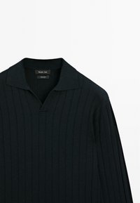 Zwarte gebreide trui met verticale ribbels, voorzien van een opstaand kraag en een halve knopenlijst. Materiaallabel zichtbaar bij de halslijn.