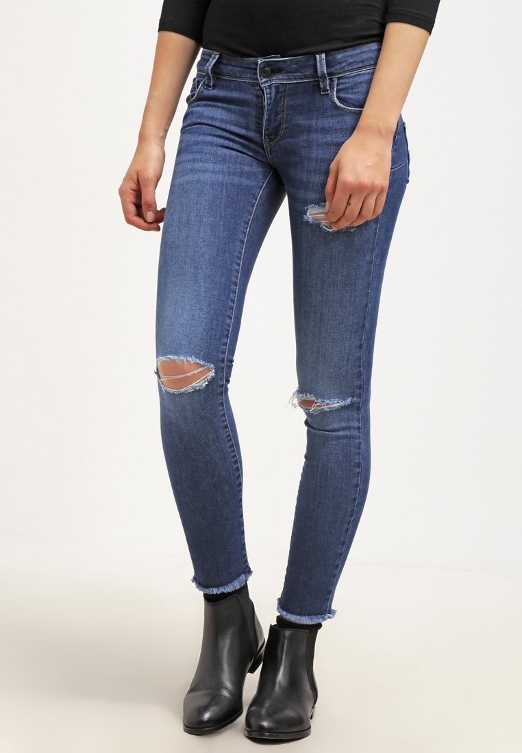 Skinny jeans in medium wash blue denim, featuring ripped knee details, a frayed hem, and a classic five-pocket design. Paired with black ankle boots.