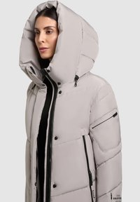 Kvinde iført en lys grå puffjakke med en stor hætte, sort lynlås og lommer, der står med venstre side mod et neutralt baggrund.