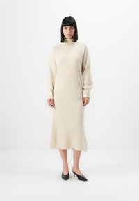 Donna in piedi che indossa un abito midi lavorato a maglia di colore cremisi a maniche lunghe con colletto alto e tacchi kitten neri a punta su uno sfondo bianco.