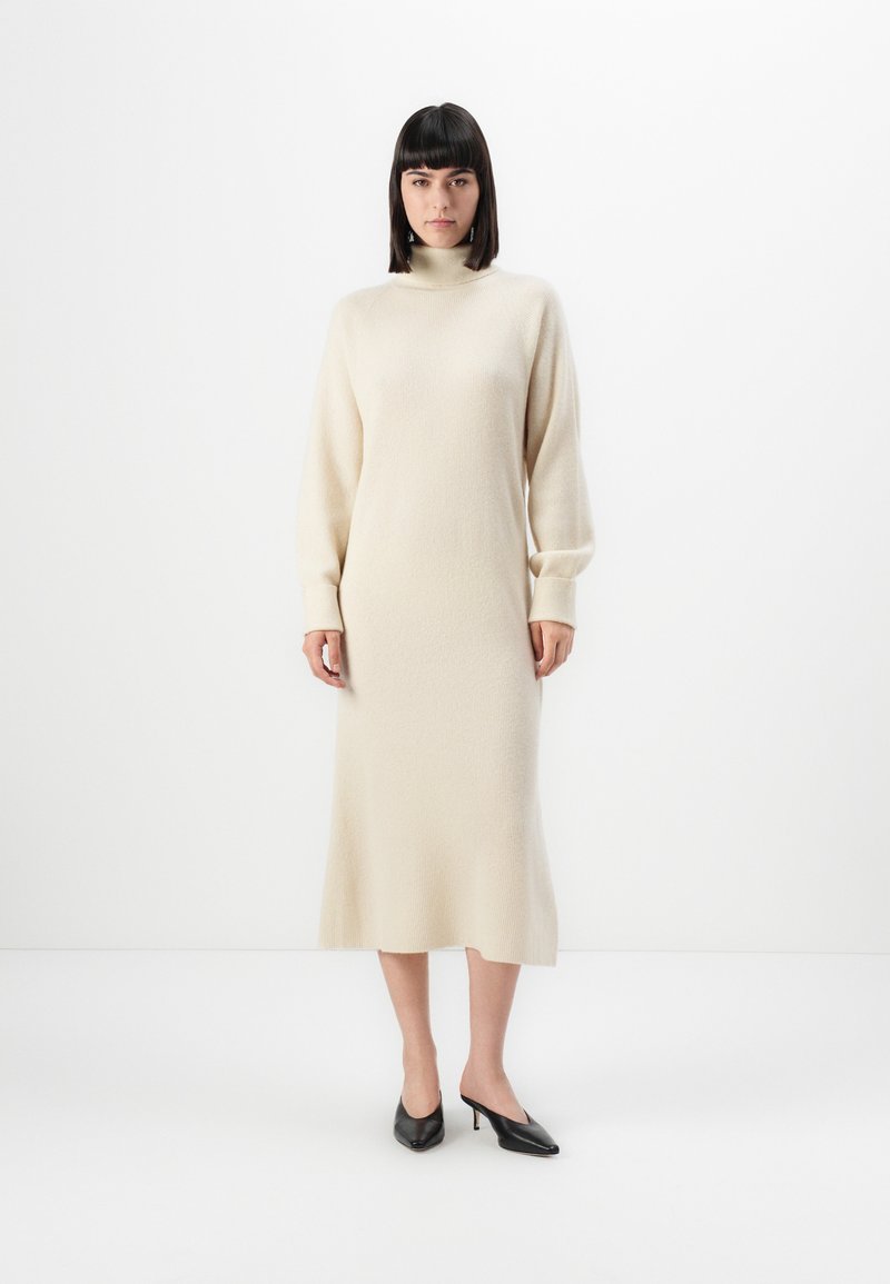 Uma mulher está de pé vestindo um vestido midi de malha de manga longa em tom creme, com gola alta, e sapatos de salto kitten heels pretos, em um fundo branco.