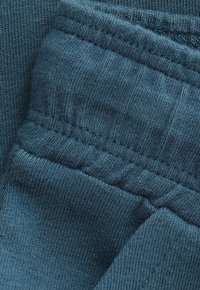 Zbliżenie na turkusową tkaninę ukazującą miękką, prążkowaną teksturę dzianiny oraz stębnowanie elastycznego pasa na złożonym ubraniu.