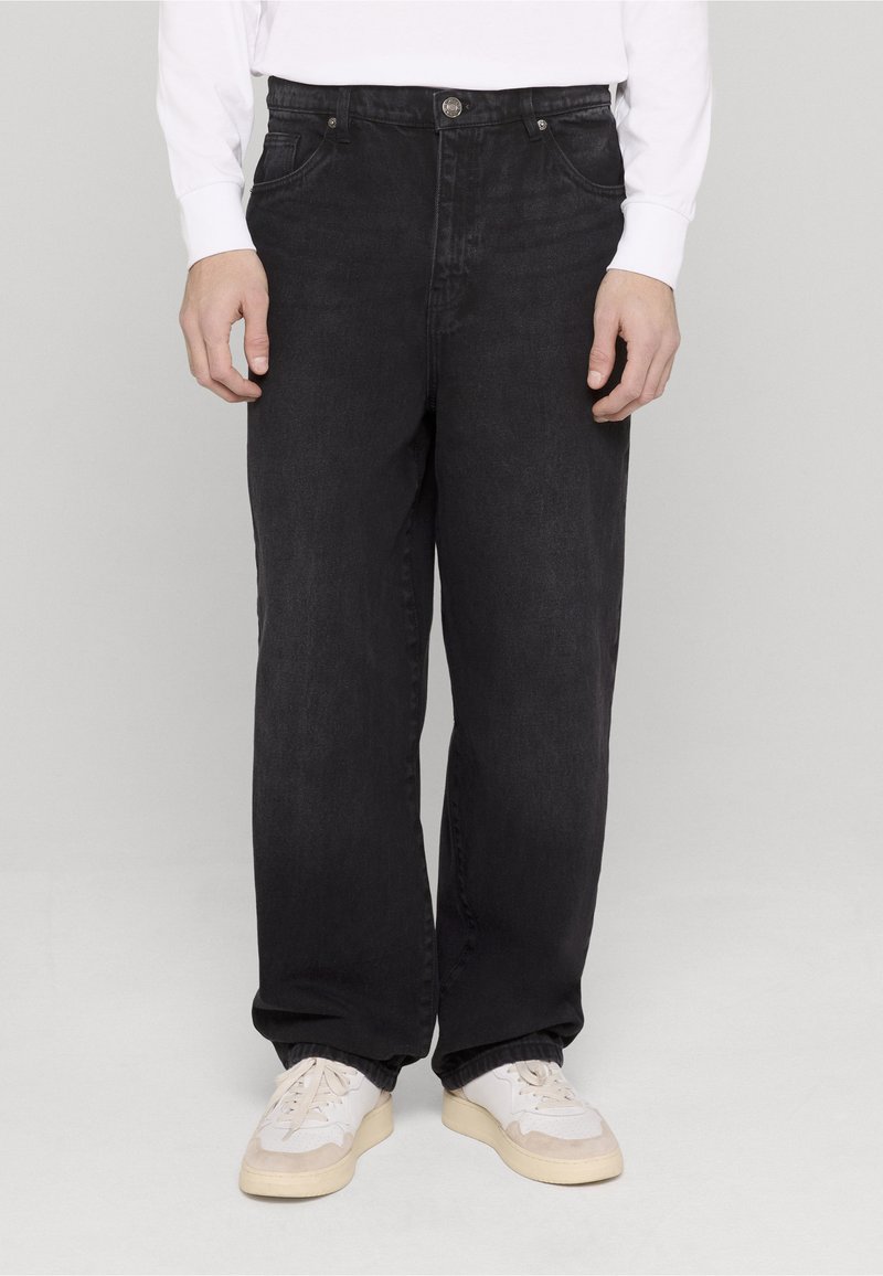 Mustad sirge lõikega teksapüksid, millel on kõrge vöökoht, viis taskut ja ees nööpkinnitusega sulgumine. kantud valgete tossudega.