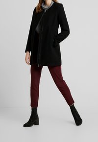 Vrouw in een zwarte ritsjas, gestreepte grijze top, bordeauxrode broek en zwarte enkellaarsjes met hak, staande tegen een effen grijze achtergrond.