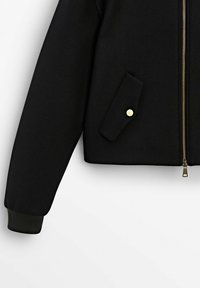 Veste noire à manches longues avec poignets côtelés, fermeture éclair devant et poche à rabat sécurisée par un bouton doré sur le côté inférieur.