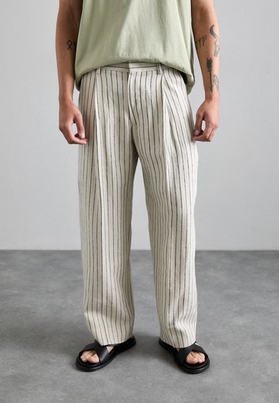 Helle beige gestreifte Hosen mit lockerem Schnitt und Falten, aus einem strukturierten Stoff gefertigt. Getragen mit schwarzen Sandalen, die ein minimalistisches Design aufweisen.