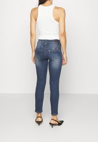 Jeans ajustados en denim de azul medio, con una cintura alta, detalles desgastados y dos bolsillos traseros con cremalleras. Combinados con una camiseta sin mangas blanca.