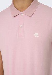 Camisa polo rosa claro con una tela texturizada, tres botones en el cuello y un pequeño logotipo blanco en el lado izquierdo del pecho.