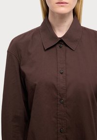 Camisa marrón de botones, hecha de una tela suave, con cuello puntiagudo y botones negros en el frente, y mangas largas.