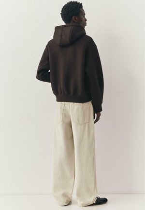 Homme portant un sweat à capuche marron foncé, un jean large écru et des baskets noires, debout face à un mur uni de couleur claire.