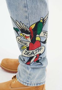 Světle modré džíny z denimu s barevným vyšitým designem ptáka, s prapory "PRAVDA" a "SMRT", doplněné hnědými botami.