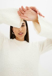 Maglione color crema con un tessuto a maglia testurizzato, collo alto e ampie maniche a campana. Il modello solleva le braccia, mostrando il design delle maniche.